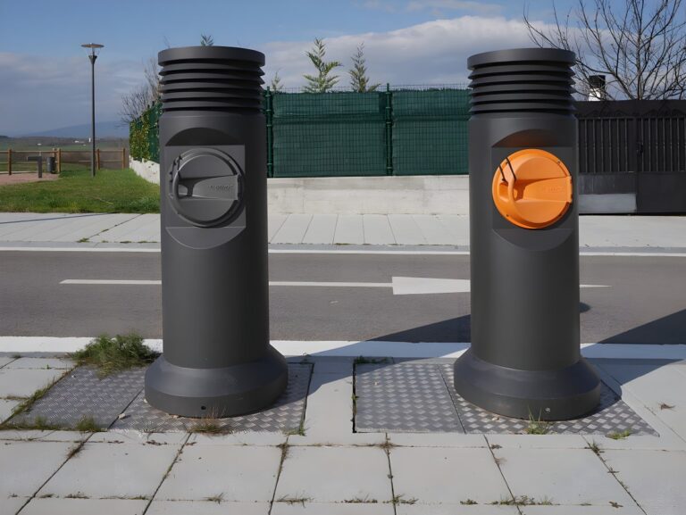 Two cylindrical pneumatic waste collection inlets on a sidewalk next to a paved road with a white directional arrow.