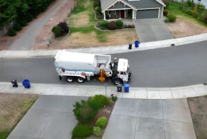 Aerial view of a white side-loader recycling truck collecting blue bins on a suburban residential street.