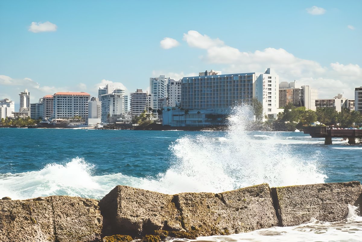 Ocean waves crashing against a stone seawall with the San Juan, Puerto Rico skyline and hotels in the background.