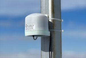 A PurpleAir air quality sensor mounted to a metal utility pole with zip ties against a blue sky.