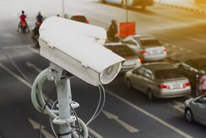 White traffic surveillance cameras mounted on a pole overlooking a busy multi-lane city road with cars and motorcycles.