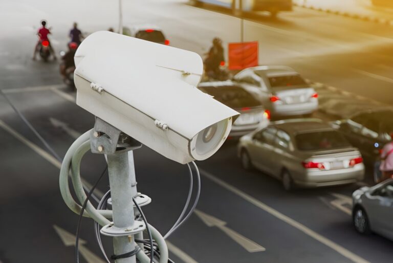 White traffic surveillance cameras mounted on a pole overlooking a busy multi-lane city road with cars and motorcycles.