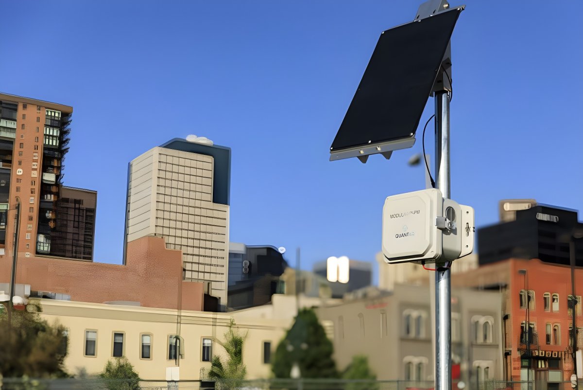 Solar-powered QuantAQ air quality sensor mounted on a metal pole with a city skyline in the background.
