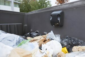 A black IoT waste sensor mounted to the interior wall of a dumpster filled with trash bags and cardboard.