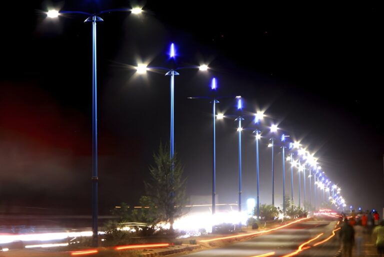 A row of illuminated Ragni Group street lamps with blue finials lining a highway at night with light trails from traffic.