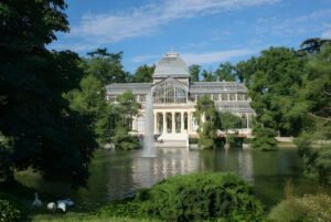 The Crystal Palace in Madrid's Retiro Park viewed across a lake with a central water fountain and surrounding trees.