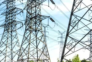 Steel lattice transmission towers and high-voltage power lines against a bright blue sky.