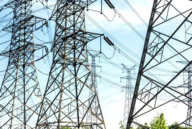 Steel lattice transmission towers and high-voltage power lines against a bright blue sky.