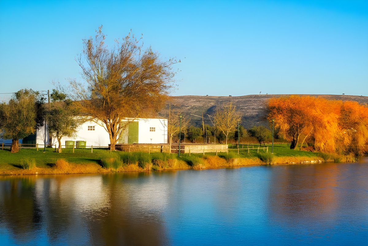 Small white utility building on a grassy bank beside a rural river with autumn trees and hills in the background.