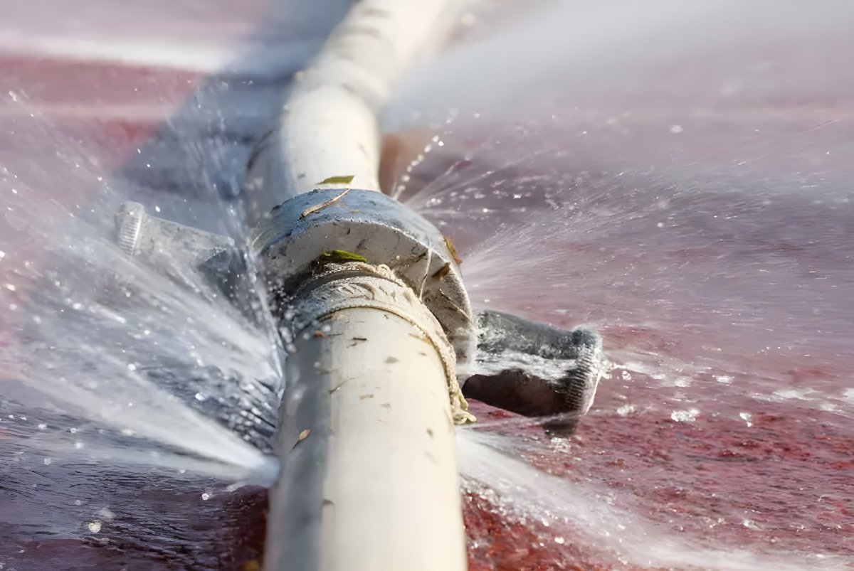 Water spraying from multiple leaks in a white industrial pipe over a red surface.