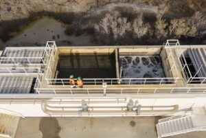 Aerial view of two workers in safety vests standing on a walkway above industrial wastewater treatment aeration tanks.