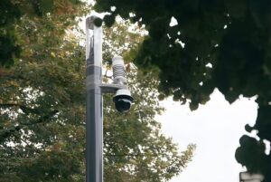Schréder Shuffle smart pole with integrated security camera and environmental sensors positioned among green trees.