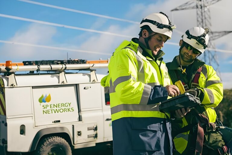 Two SP Energy Networks technicians in high-visibility gear use a tablet near a utility van and power lines.