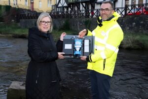 Two people hold a Scottish Water sewer monitor device in front of a river and stone wall.