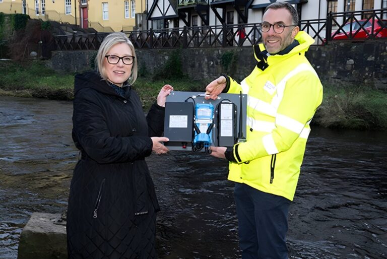 Two people hold a Scottish Water sewer monitor device in front of a river and stone wall.