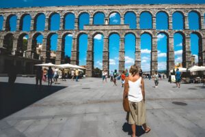 Pedestrians walk through a stone plaza in front of the ancient Roman Aqueduct of Segovia under a clear blue sky.