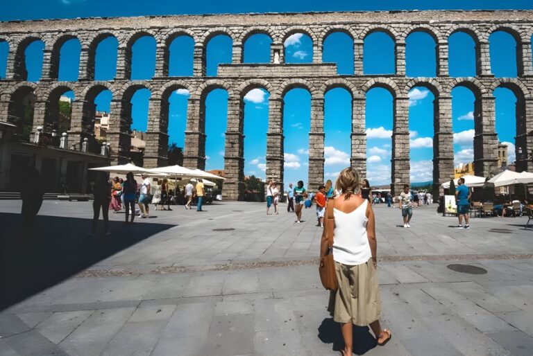 Pedestrians walk through a stone plaza in front of the ancient Roman Aqueduct of Segovia under a clear blue sky.