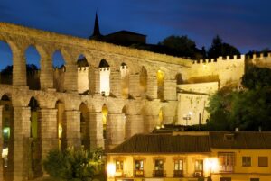 The Roman Aqueduct of Segovia and surrounding historic buildings illuminated by warm street lighting at night.
