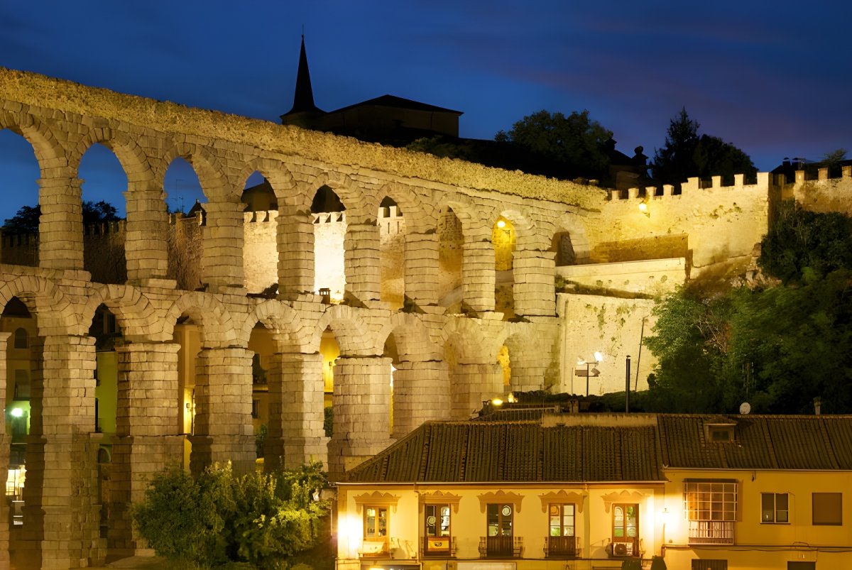 The Roman Aqueduct of Segovia and surrounding historic buildings illuminated by warm street lighting at night.