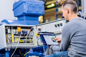 A technician uses a tablet to monitor data next to an open electrical control panel on industrial machinery.