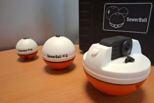 Three Suez SewerBall monitoring devices in various sizes on a wooden surface next to a branded black carrying case.