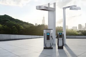 Two Siemens eMobility electric vehicle charging stations and overhead pantographs on a paved lot with a city skyline.