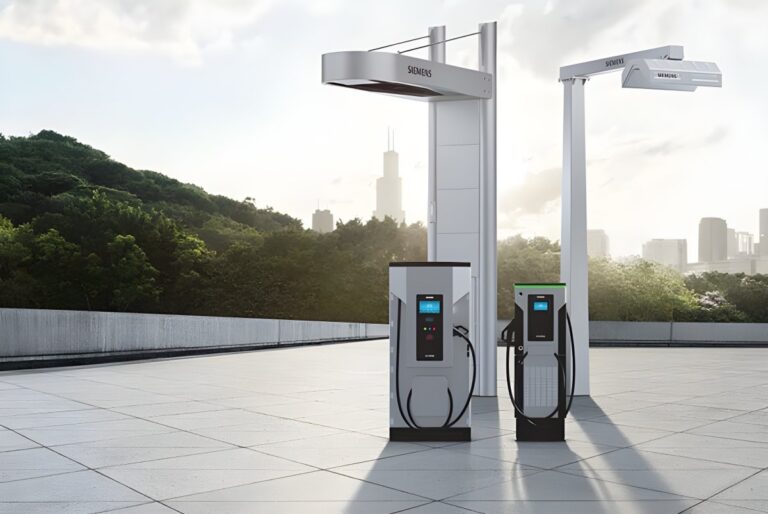 Two Siemens eMobility electric vehicle charging stations and overhead pantographs on a paved lot with a city skyline.