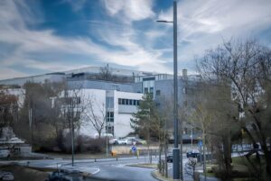 A 5G Synergiewerk smart pole stands on a street corner in front of a multi-story office building and parking lot.