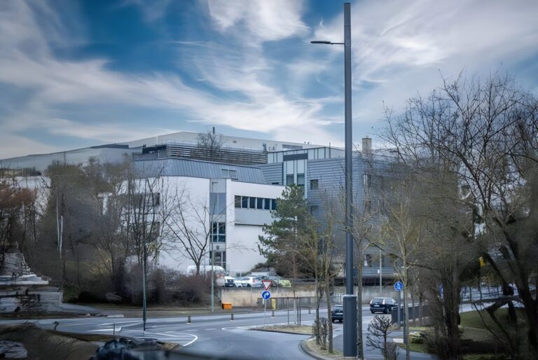A 5G Synergiewerk smart pole stands on a street corner in front of a multi-story office building and parking lot.