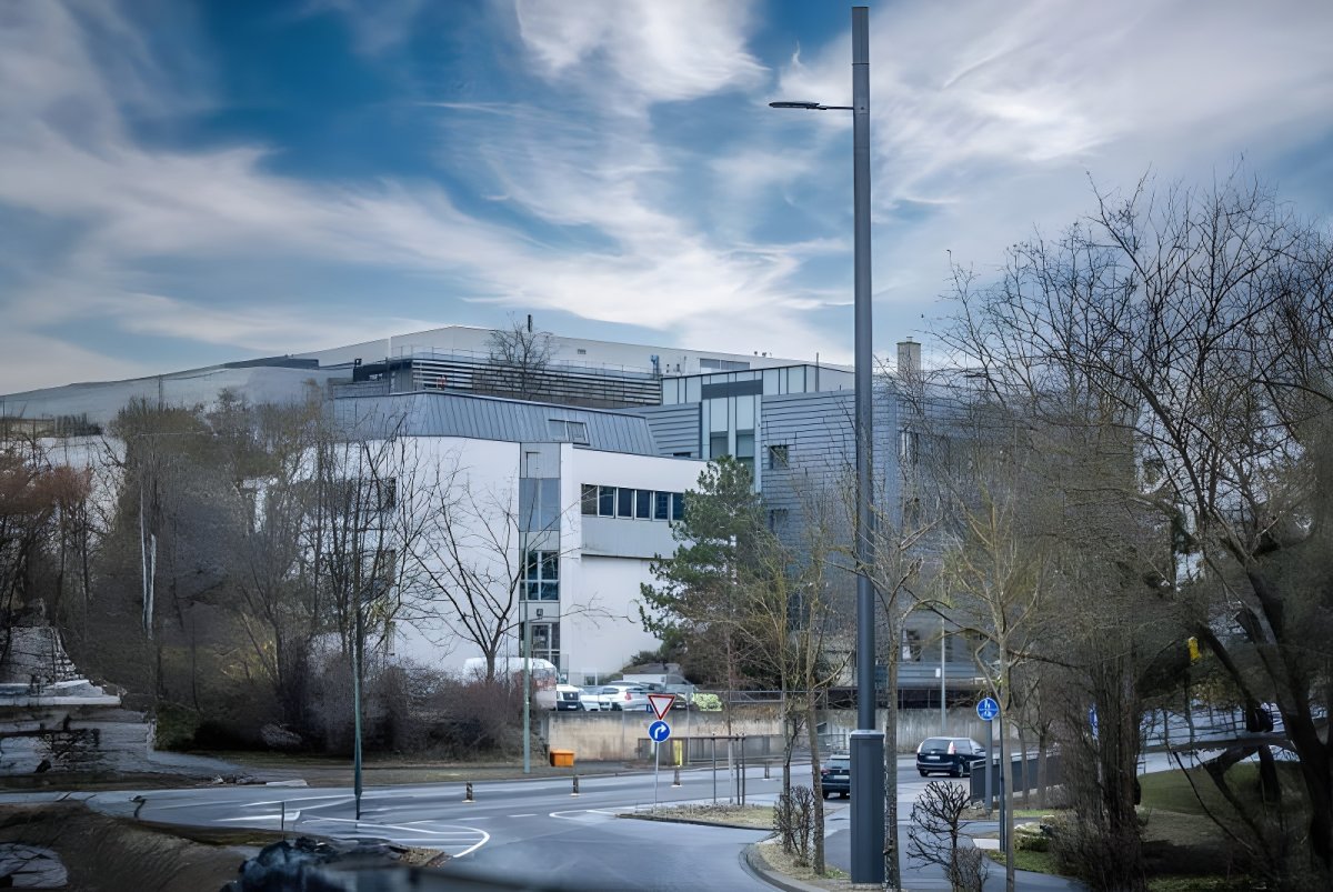 A 5G Synergiewerk smart pole stands on a street corner in front of a multi-story office building and parking lot.