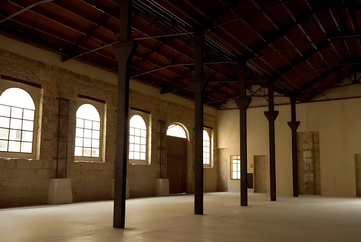 Interior of an empty industrial warehouse with stone walls, arched windows, and dark steel support columns.