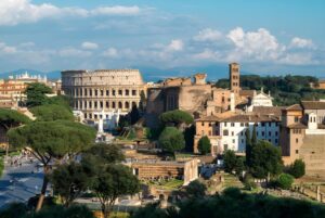 Panoramic view of the Roman Colosseum and surrounding ancient ruins under a blue sky with scattered clouds.