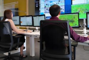 Two operators in a control room monitoring digital maps and data on multiple computer screens and a large wall display.