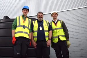 Three workers in high-visibility vests and hard hats stand in front of a large black water tank and a brick building.