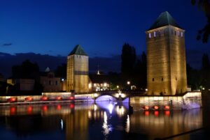 Illuminated medieval towers and the Ponts Couverts bridge reflected in the Ill River at night in Strasbourg, France.