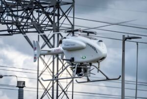 A white SwissDrones SDO 50 V2 unmanned helicopter flying in front of high-voltage power lines and utility infrastructure.