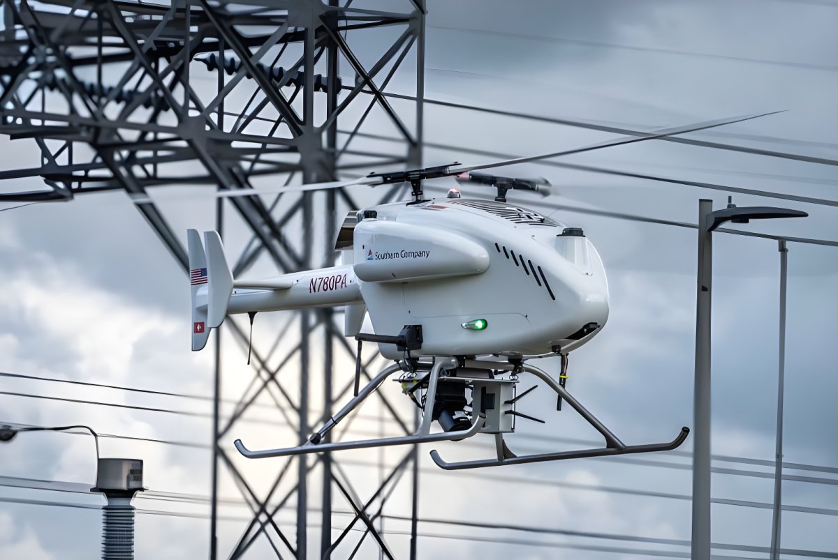 A white SwissDrones SDO 50 V2 unmanned helicopter flying in front of high-voltage power lines and utility infrastructure.