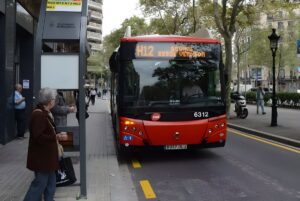 Red TMB H12 bus approaching a street-side bus stop with waiting passengers on a Barcelona sidewalk.
