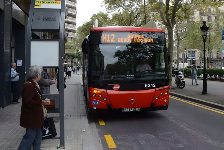 Red TMB H12 bus approaching a street-side bus stop with waiting passengers on a Barcelona sidewalk.