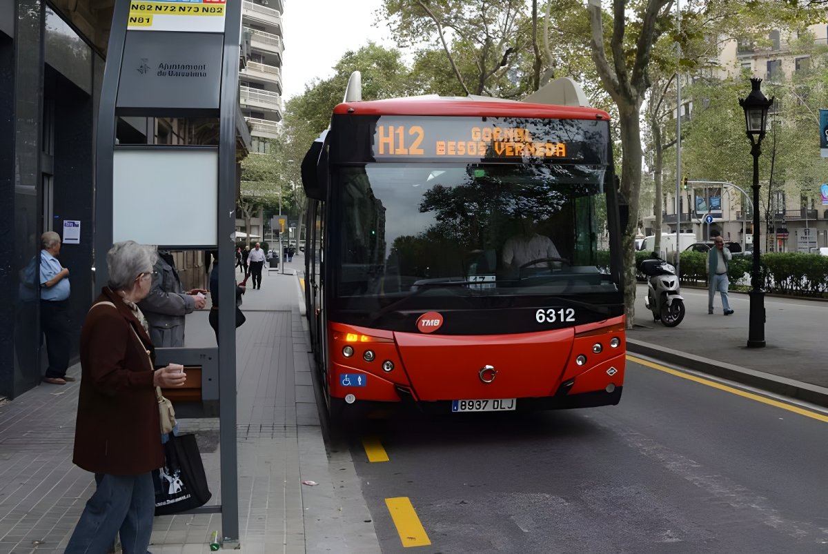 Red TMB H12 bus approaching a street-side bus stop with waiting passengers on a Barcelona sidewalk.