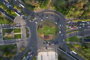 Top-down aerial view of a busy multi-lane urban traffic roundabout with cars and surrounding green trees.