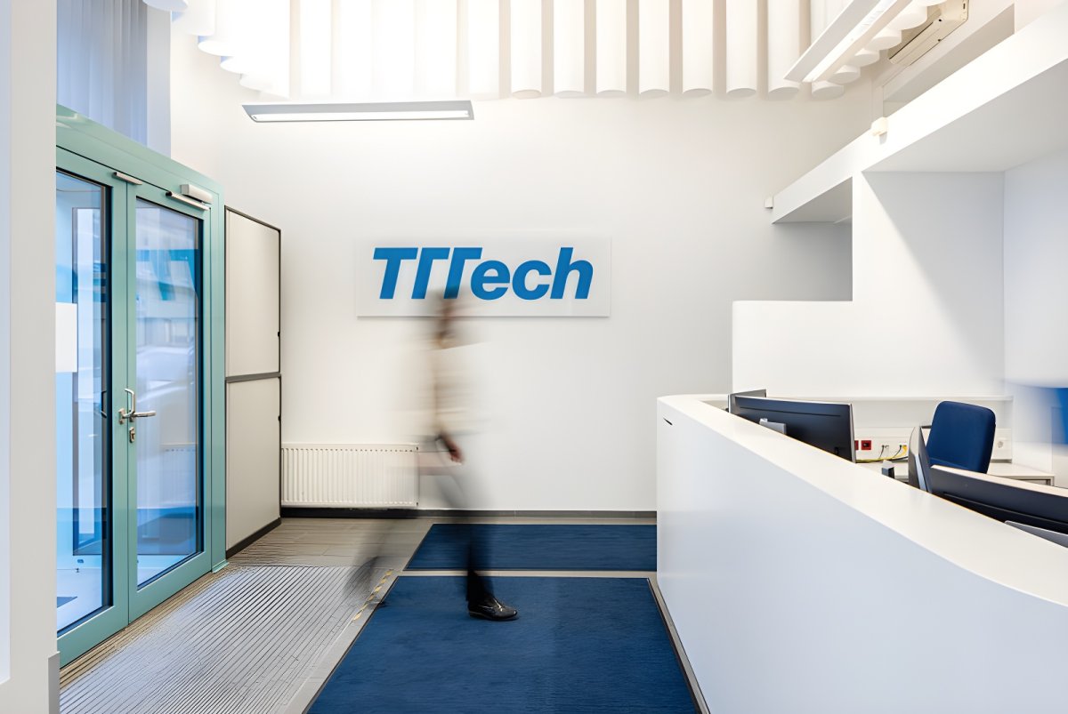 A blurred person walks past a white reception desk toward a wall featuring the TTTech logo in a bright office lobby.