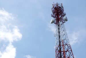 Red and white lattice telecommunications tower with satellite dishes against a blue sky with light clouds.