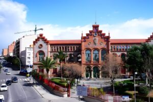The red brick facade of the Industrial School of Terrassa building viewed from a street with palm trees and traffic.