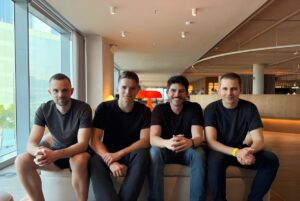 Four men in dark t-shirts sit on a bench in a modern office lounge with floor-to-ceiling windows.