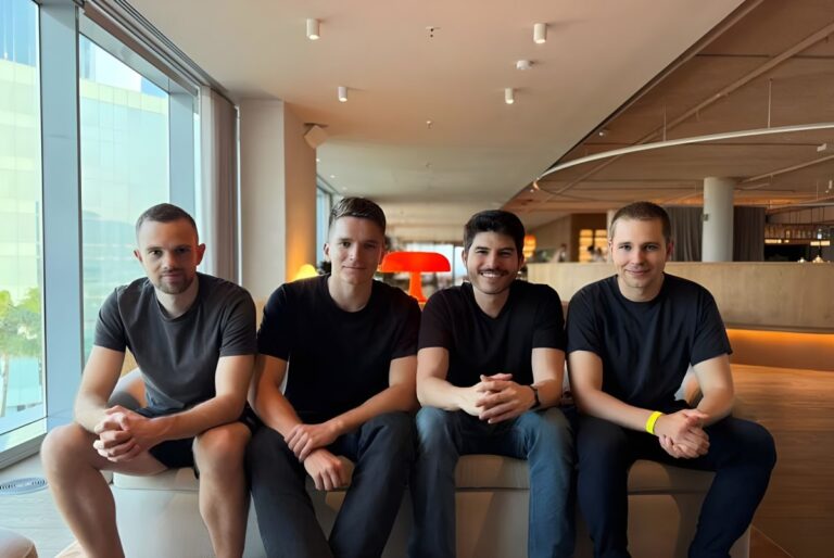 Four men in dark t-shirts sit on a bench in a modern office lounge with floor-to-ceiling windows.