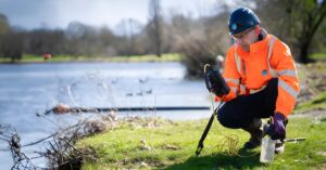 A Thames Water technician in high-visibility gear uses a handheld sensor to test water quality at a riverbank.