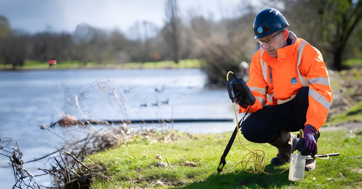 A Thames Water technician in high-visibility gear uses a handheld sensor to test water quality at a riverbank.