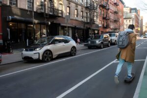 A person on a skateboard rides past a white BMW i3 parked on a city street with brick buildings and a bike lane.