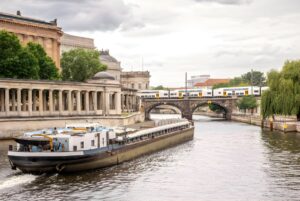 A cargo barge travels down the Spree River in Berlin past Museum Island and a train crossing a stone arch bridge.